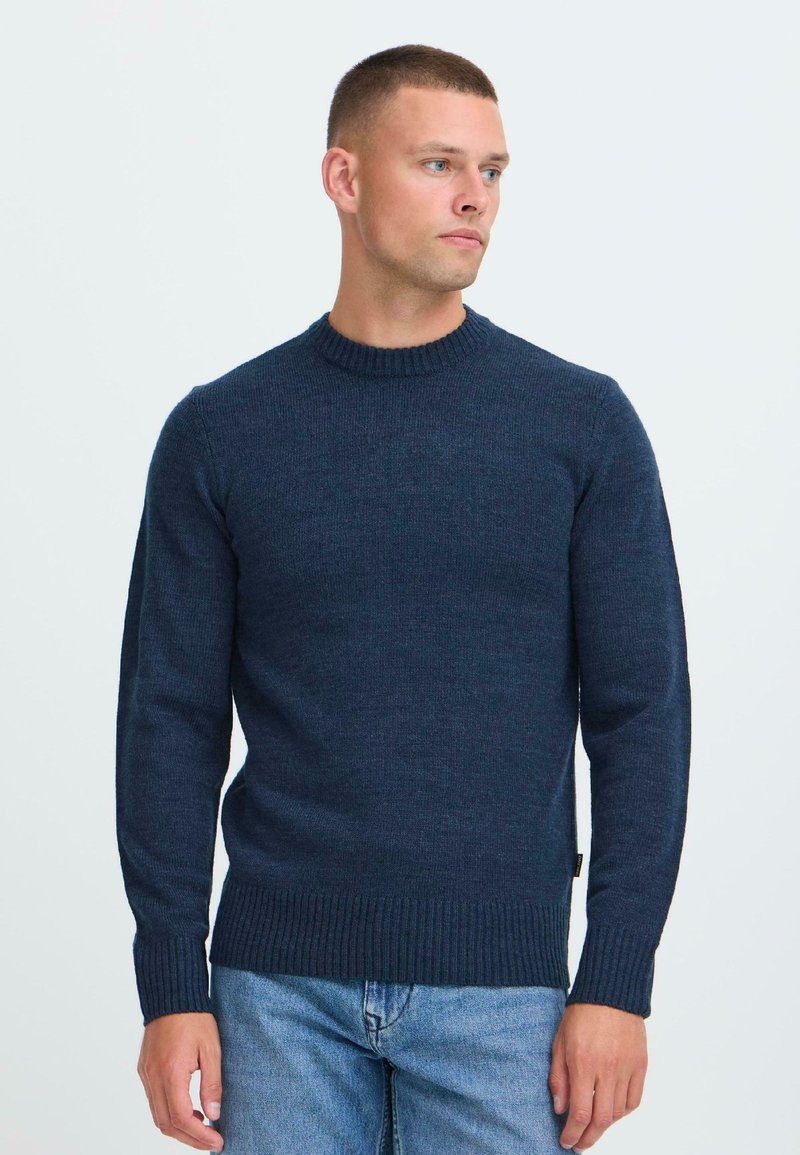 Mann med kort hår iført en marineblå strikkegenser og lyseblå jeans, som ser mot høyre mot en enkel hvit bakgrunn.