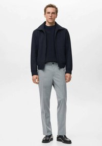 Chaqueta navy con cremallera sobre un suéter navy, combinada con pantalones rayados en gris claro y zapatos de cuero negros. Estilo casual y paleta de tonos neutros.