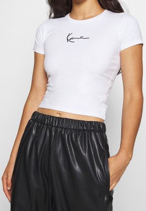 T-shirt cropped blanc ajusté avec un logo noir brodé sur la poitrine, manches courtes et une texture de tissu lisse. Associé à un pantalon noir en simili cuir.