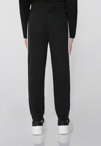 Schwarze Sweatpants mit lockerer Passform, elastischem Bund und weißen Seitenstreifen, kombiniert mit weißen Sneakern, von hinten betrachtet.