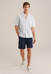 Muž jde oblečený v bílém tričku s krátkým rukávem a modrými pruhy, tmavě modrých šortkách a světle zbarvených teniskách na jednobarevném pozadí.