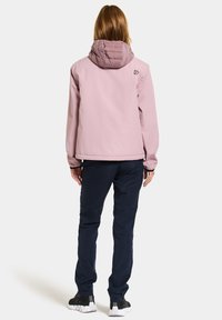 Personne avec de longs cheveux portant une veste à capuche violette claire, un pantalon foncé et des baskets noires, se tenant de dos sur un fond uni.