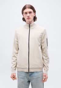 Νεαρός άνδρας με μακριά μαλλιά μέχρι τους ώμους, ντυμένος με μπεζ μπουφάν με φερμουάρ σε μαύρο χρώμα και ανοιχτό μπλε τζιν, στέκεται μπροστά σε απλό φόντο.