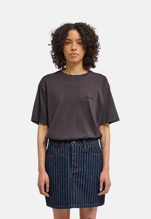Donna con capelli ricci che indossa una T-shirt oversize scura e una gonna di jeans blu a righe, in piedi davanti a uno sfondo semplice.