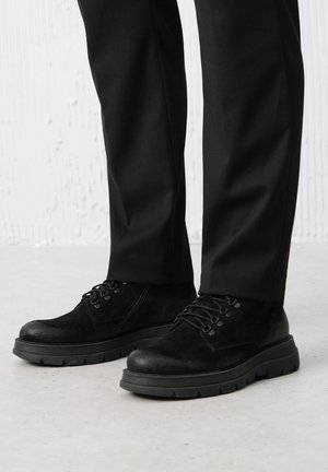Czarne zamszowe buty o teksturowanej powierzchni, z masywną gumową podeszwą, z zapięciem na sznurowadła oraz subtelnym wykończeniem ściegów.