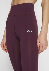 Legíny s vysokým pasem v barvě švestek vyrobené z elastického materiálu. Mají hladký povrch a malé bílé logo v oblasti boků.