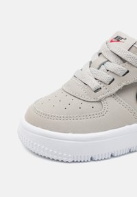 Világosszürke edzőcipő sima velúr textúrával, perforált orr résszel, lapos fűzőkkel és csillag alakú talppal rendelkező fehér gumitalppal. A Nike logó látható.
