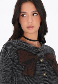 Cardigan tricotat în nuanțe de negru și gri, cu un material texturat. Include aplicații mari sub formă de fundițe maro și butoane în nuanță aurie pe partea din față.