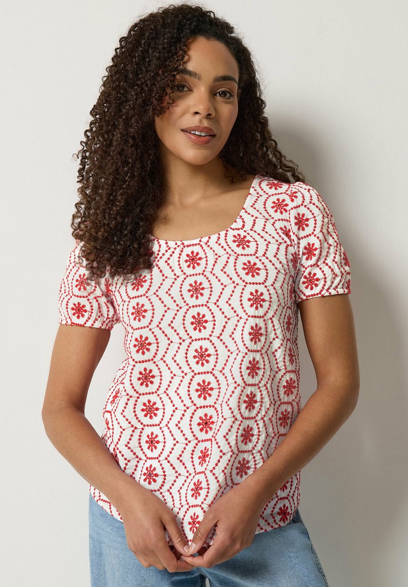 Femme aux cheveux bouclés portant un haut blanc à manches courtes avec des motifs floraux rouges et un jean bleu clair, sur un fond uni.