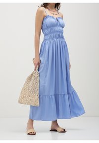 Robe longue bleu clair sans manches avec corsage froncé et jupe à volants. Le mannequin tient un sac tissé beige et porte des sandales plates marron clair.