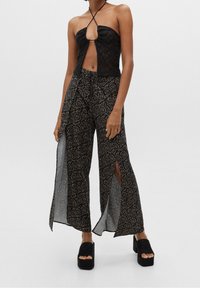 Femme portant un haut en dentelle noire à halter et un pantalon large noir avec des motifs floraux beiges et de hautes fentes, associé à des sandales à plateforme noires.