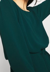 Turkusowa sukienka z długim rękawem, o kwadratowym dekolcie, miękkiej fakturze i związanym w talii detalu. Materiał wydaje się lekki i lekko zwiewny.