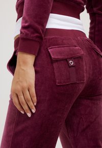 Burgundy velour joggers with a textured finish, featuring side pockets and a fitted waistband with contrasting white stripe.