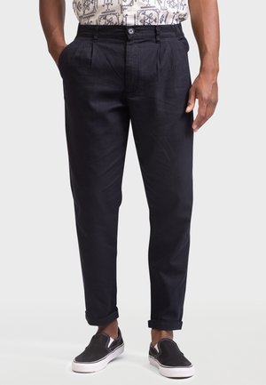 Mann trägt schwarze, schmal geschnittene Hosen mit umgeschlagenen Säumen, ein gemustertes weißes Hemd, das hineingesteckt ist, und schwarze Slip-On-Sneaker mit weißen Sohlen.