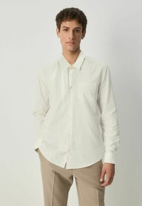 Cămașă lungă cu mânecă, de culoare crem deschis, cu guler ascuțit, imprimeu cu puncte mici, buzunar frontal și închidere cu nasturi, combinată cu pantaloni bej.