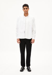 Witte blouse met lange mouwen en een button-down kraag, met een enkele borstzak, gecombineerd met zwarte broek en zwarte schoenen, staande op een witte achtergrond.