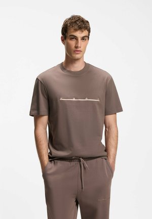 Ung mand iført en brun kortærmet t-shirt og matchende bukser med hænderne i lommerne mod en ensfarvet hvid baggrund.