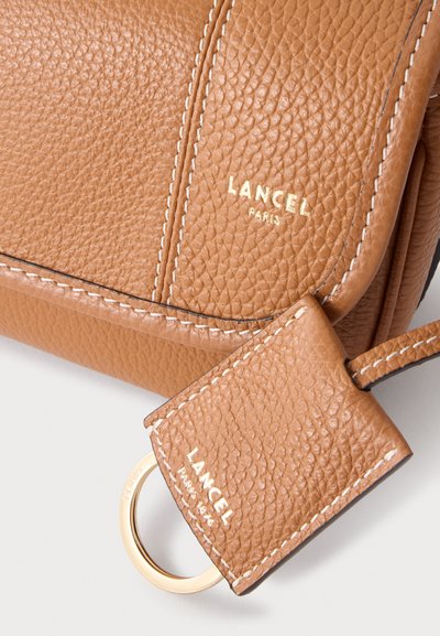 Кадър отблизо на чанта Lancel Paris от кафява шагренова кожа, с бели шевове, кожен езичок на каишката с марка и златист пръстен.