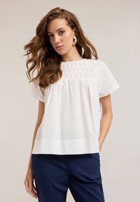 Blusa blanca con mangas cortas, que presenta un detalle de yoke texturizado y un corte holgado. Combinada con pantalones azul marino para crear contraste.