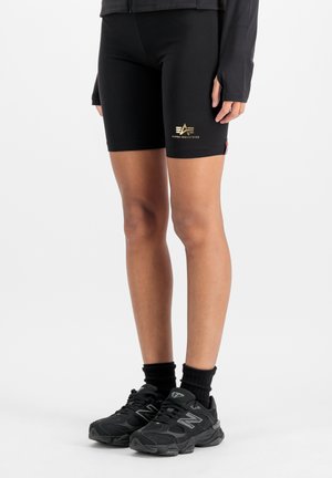 Nogi w czarnych krótkich spodenkach motocyklowych Alpha Industries z złotym logo, czarne skarpetki i czarne sneakersy New Balance na białym tle.