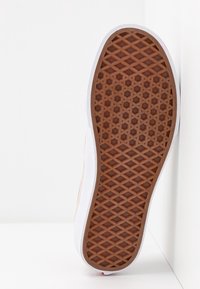 Suola di scarpa con battistrada in gomma marrone a motivo waffle e intersuola bianca, posizionata verticalmente contro uno sfondo angolare bianco.