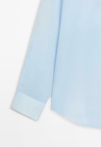 Chemise bleu clair à manches longues avec un poignet visible sur un fond blanc uni.
