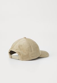 Beige cordfløyelslue med buet skjerm og justerbar stropp bak. Har teksturert ribbing og ventilasjonslufthull.