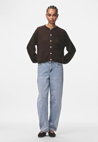 Rjav cardigan z belimi gumbi, sproščen kroj, dolgi rokavi, nošen nad svetlomodrimi ravnimi hlačami, v kombinaciji s črnimi čevlji na zdrs.