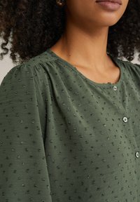Blusa verde con texture, caratterizzata da un collo rotondo, piccoli bottoni e un design arricciato con sottili punti decorativi sparsi sulla stoffa.