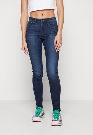 Dark blue skinny jeans with classic five-pocket design, featuring a front button and zipper closure. Worn with green and pink sneakers.