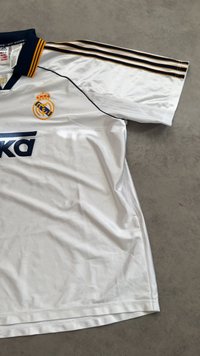 Bílý fotbalový dres Real Madrid se námořnickým a zlatým pruhovaným límcem, klubovým znakem na hrudi, značkou Adidas a logem sponzora na přední straně.