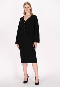 Schwarze Strickjacke mit V-Ausschnitt, drei goldenen Knöpfen vorne, kombiniert mit einem knielangen Rock aus demselben Material und in derselben Farbe.