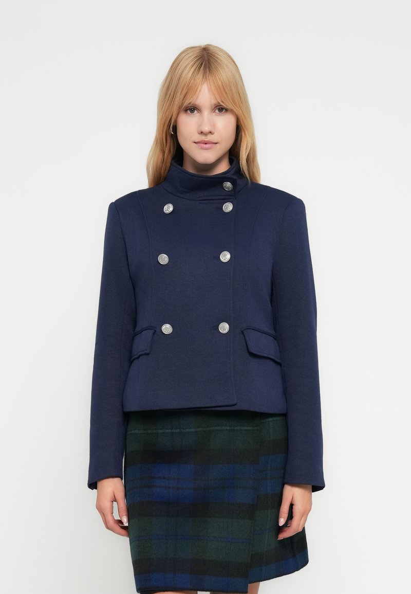 Μπλε διπλόπιετο σακάκι με ψηλό γιακά, ασημένια κουμπιά, τσέπες στα πλάγια και υφή, φορεμένο πάνω από καρό φούστα.