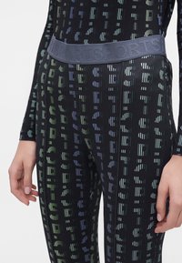 Gros plan d'une personne portant des leggings noirs et un haut avec des lettres imprimées répétées et une large ceinture élastique portant la mention "SPORTS."