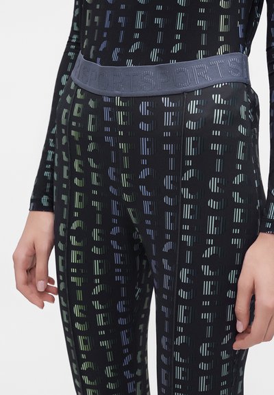 Gros plan d'une personne portant des leggings noirs et un haut avec des lettres imprimées répétées et une large ceinture élastique portant la mention "SPORTS."