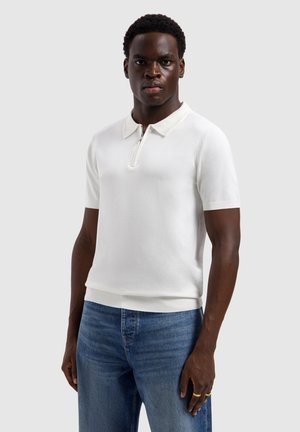 Jonge man die een wit poloshirt met korte mouwen en een ritssluiting aan de voorkant draagt en een blauwe spijkerbroek, staand tegen een effen lichte achtergrond.