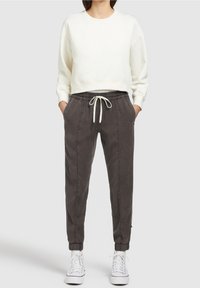 Vrouw in een witte cropped sweatshirt, donkergrijze joggingsbroek met trekkoord en boorden, en witte hoge sneakers, staand met de handen in de zakken.