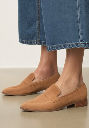 Zamszowe mokasyny w kolorze jasnobrązowym noszone z krótkimi niebieskimi jeansami na gładkiej beżowej podłodze.