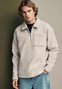 Pullover in pile grigia chiara con colletto a mezza zip, tasca frontale e superficie testurizzata, abbinato a jeans in denim blu.