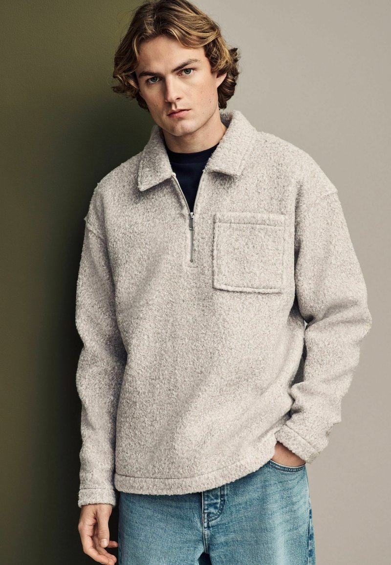Pullover in pile grigia chiara con colletto a mezza zip, tasca frontale e superficie testurizzata, abbinato a jeans in denim blu.