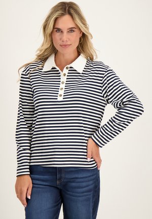 Vrouw draagt een marineblauw-wit gestreept shirt met lange mouwen, een witte kraag en knopen, gecombineerd met donkerblauwe jeans.