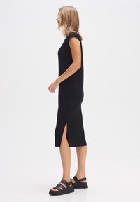 Zwarte geribbelde midi-jurk met korte mouwen en zijsplitsingen, gecombineerd met zwarte sandaaltjes met banden. Vloeiende textuur en gestroomlijnde silhouet.
