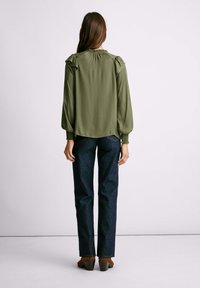 Blusa verde oliva con spalle arricciate e maniche testurizzate, abbinata a jeans in denim scuri e scarpe con tacco marroni. Sfondo semplice.