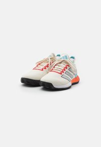 Sportowe buty z kremową cholewką, czerwonymi akcentami i szarymi paskami. Posiadają beżowe sznurówki, lekką konstrukcję i teksturowaną czarną podeszwę.
