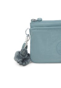 Teal nylon pung med en sølvfarvet lynlås, udstyret med en pelsagtig gorilla nøglekæde og et cirkulært logo patch på fronten.