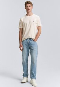 Bež bombažna majica z majhnim zelenim logotipom, kombinirana s svetlo modrimi kavbojkami iz jeansa. Obleko dopolnjujejo beli športni copati.