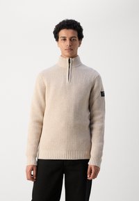 Suéter de punto acanalado beige con cuello alto y cierre de media cremallera. Presenta una tela texturizada y un pequeño parche con el logotipo en la manga izquierda.