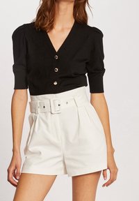 Schwarze gestrickte Strickjacke mit V-Ausschnitt, kurzen Puffärmeln und drei metallischen Knöpfen, kombiniert mit weißen Shorts in hoher Taille mit einem Gürtel.