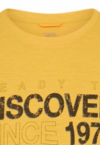 Gelbes T-Shirt mit Rundhalsausschnitt und abgenutztem schwarzen und blauen Text, auf dem "DISCOVER" sowie teilweise "READY T" und "SINCE 197" zu sehen sind.