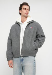 Graue Fleecejacke mit Stehkragen, Reißverschluss vorne und gesticktem Markenlogo auf der Brust. Getragen über einem weißen Hoodie. Lässiger Schnitt.